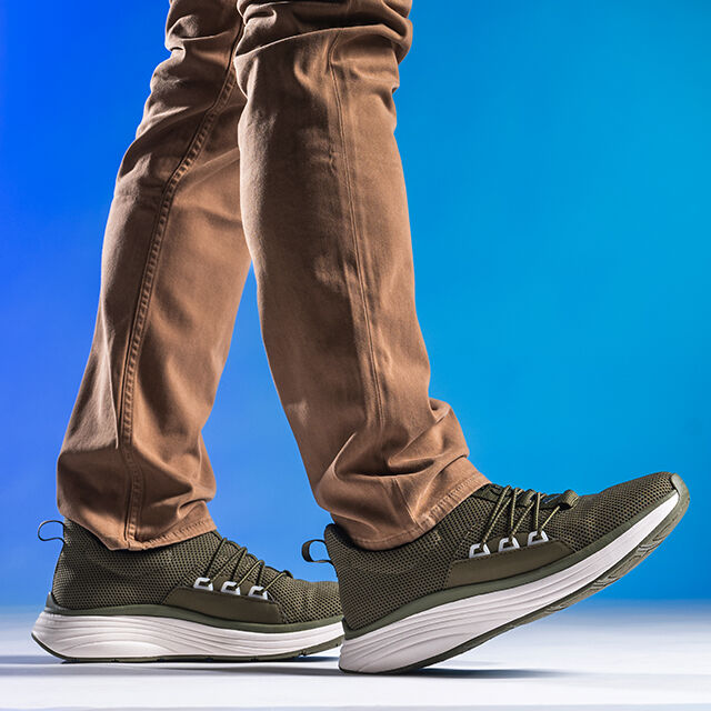 Man wearing Align Talmon in Olive with brown pants on a blue gradient background. Front foot has toe in the air.